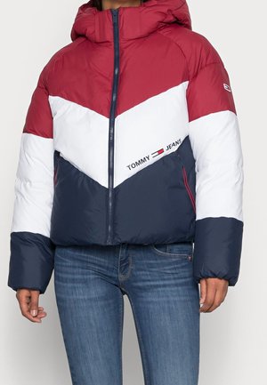 Žena nosící prošívanou bundu s barevnými bloky v červené, bílé a námořnické modři s logem „Tommy Jeans“ na přední straně, doplněnou modrými džínami.