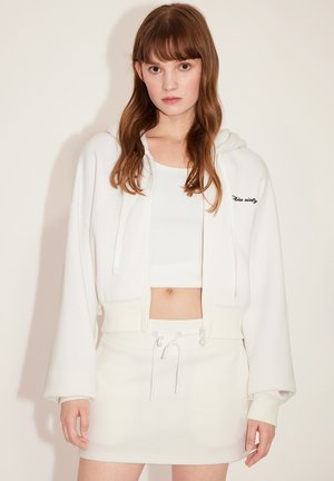 Sudadera blanca corta con cordón y texto bordado; combinada con una falda mini blanca con cordón ajustable. Tejido suave, corte relajado.