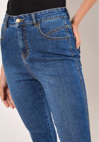 Vaqueros de mezclilla azul con un corte ajustado, diseño de cinco bolsillos, costuras visibles y cintura de altura media. La textura parece suave y lisa.