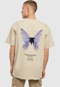 Beige oversized T-shirt med et stort lilla sommerfuglegrafik på ryggen, med sort tekst og abstrakte elementer nedenunder.