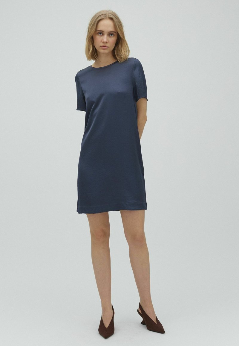 Femme aux cheveux blonds portant une robe bleu marine à manches courtes et des talons pointus marron foncé, debout devant un fond clair uni.