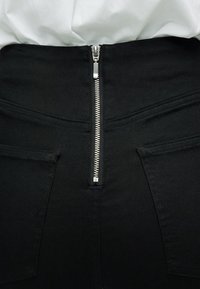 Pantalon noir avec une fermeture éclair argentée visible à l'arrière de la taille, assorti à une chemise blanche rentrée.