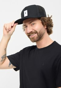 Gorra negra con visera plana y parte trasera de malla, que presenta un parche blanco con un diseño de árbol. El modelo lleva una camiseta negra sencilla con un pequeño logo.