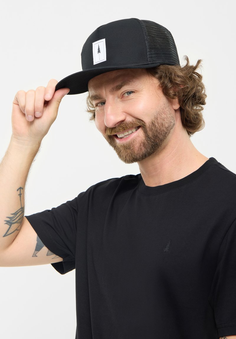 Gorra negra con visera plana y parte trasera de malla, que presenta un parche blanco con un diseño de árbol. El modelo lleva una camiseta negra sencilla con un pequeño logo.