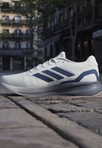 Weißer und marineblauer Laufschuh mit drei Streifen auf einer Kopfsteinpflasterstraße in einer urbanen Umgebung.
