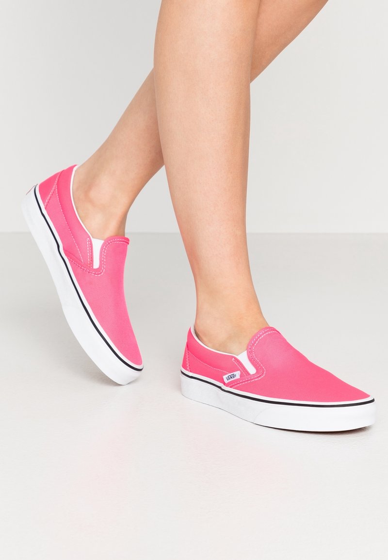 Jambes portant des baskets Vans slip-on rose vif avec semelles blanches et garniture noire, debout sur un sol clair uni.