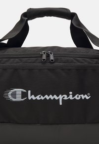 Borsa a tracolla nera con doppi zip e una maniglia imbottita. Presenta un grande logo bianco "Champion" con una "C" stilizzata sul davanti.