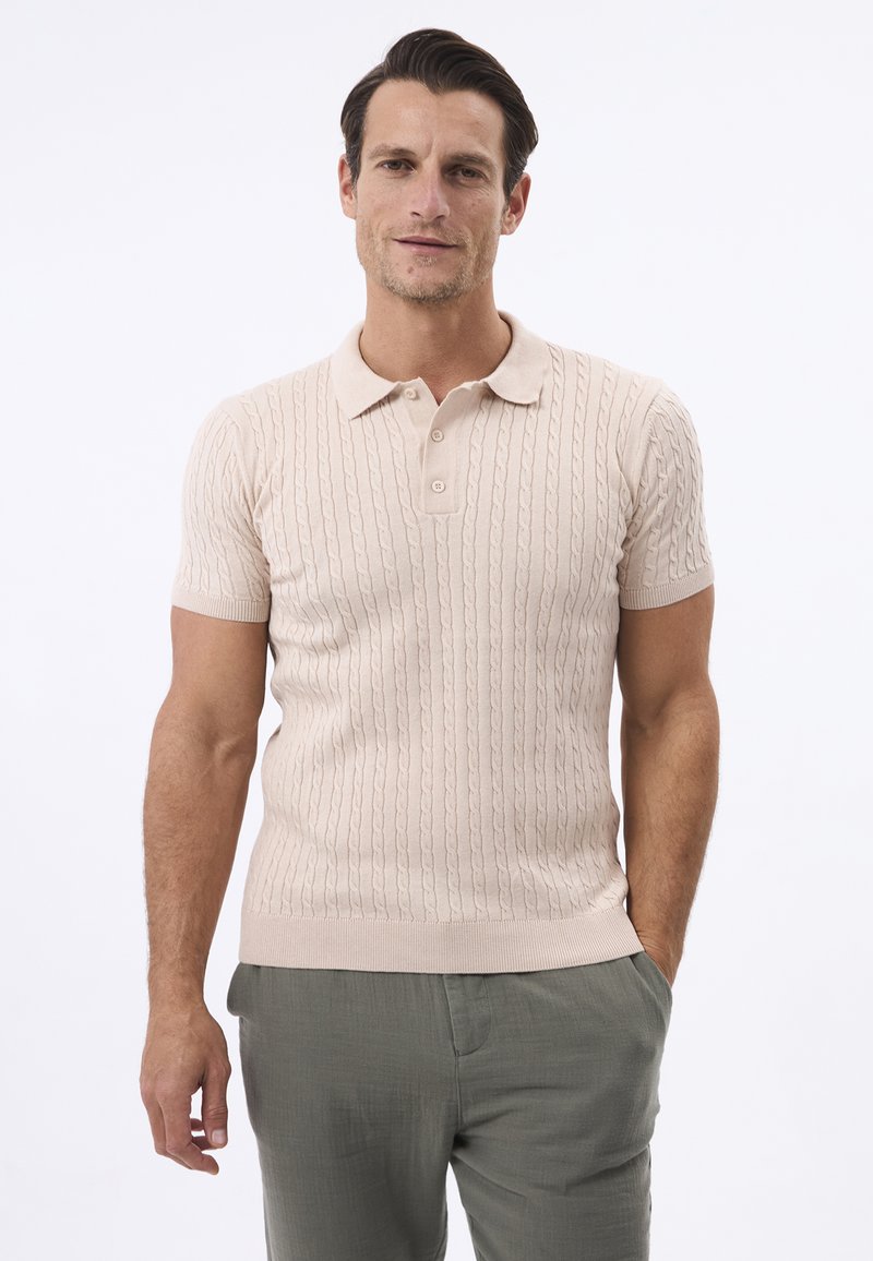 Camisa polo de manga corta en color beige claro, con textura de punto de cable, cuello y tapeta de tres botones, combinada con pantalones verdes claros.