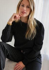 Zwart gestreept pak bestaande uit een lange mouw top en bijpassende broek, gemaakt van een gladde stof. Accenten zijn onder andere gouden sieraden en een zwarte riem.