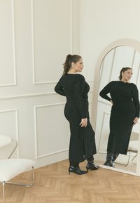 Robe noire à manches longues avec un motif texturé et une fente sur le côté, assortie à des bottines noires à bout pointu, reflétée dans un miroir en forme d'arc.