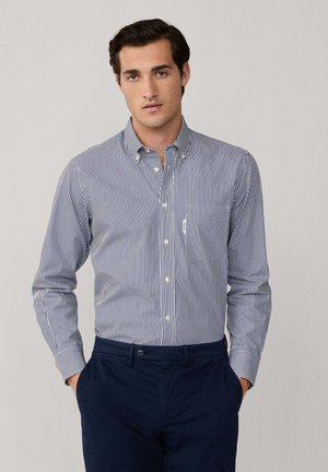 Camisa de botones a rayas azul y blanca con collar clásico, mangas largas y un bolsillo en el lado izquierdo, combinada con pantalones oscuros.