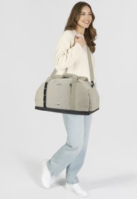 Sac de sport beige avec fond noir renforcé, double poignées, bandoulière réglable, compartiments zippés et détails en mesh.