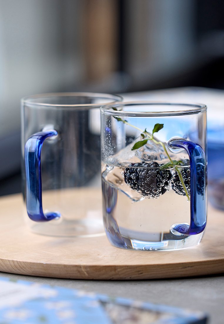 Dwa przezroczyste szklanki z niebieskimi uchwytami, wypełnione gazowaną wodą, lodem, jeżynami i gałązką zielonej mięty, na drewnianej powierzchni.