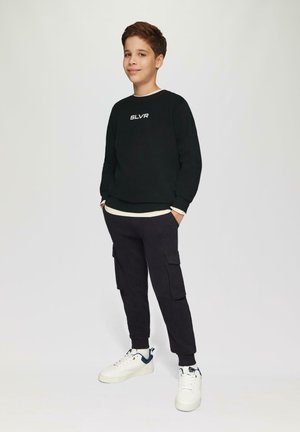 Czarna bluza z białym logo "SLVR", założona na kremową koszulę. Czarne spodnie cargo z bocznymi kieszeniami. Białe sneakersy z granatowymi akcentami.
