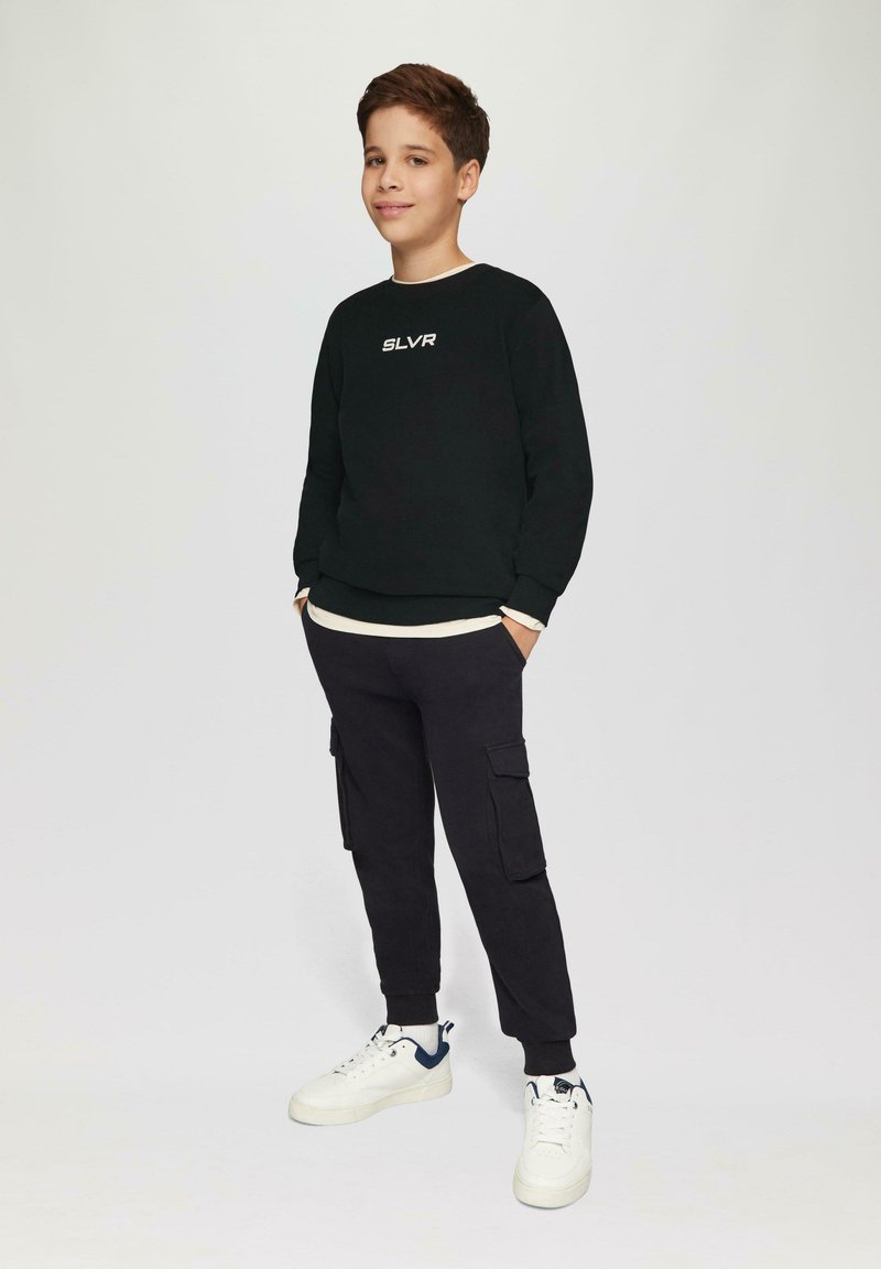 Zwarte sweatshirt met witte "SLVR" logo, gelaagd over een crèmekleurig shirt. Zwarte cargobroek met zijzakken. Witte sneakers met marineblauwe accenten.