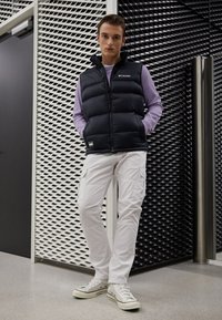 Černá péřová vesta, fialové tričko s dlouhým rukávem, bílé cargo kalhoty a světlé tenisky se zelenými akcenty. Texturované pozadí v kontrastu.