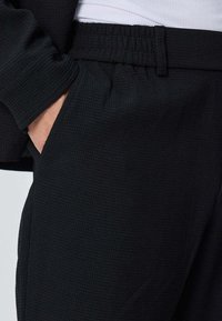 Jogging noir texturé avec une ceinture élastique et des poches latérales, présentant un motif en grille subtil pour un détail supplémentaire.