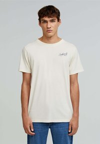 Cremefarvet bomulds t-shirt med korte ærmer, der har et lille marineblåt logo på venstre bryst og en klassisk rund halsudskæring.