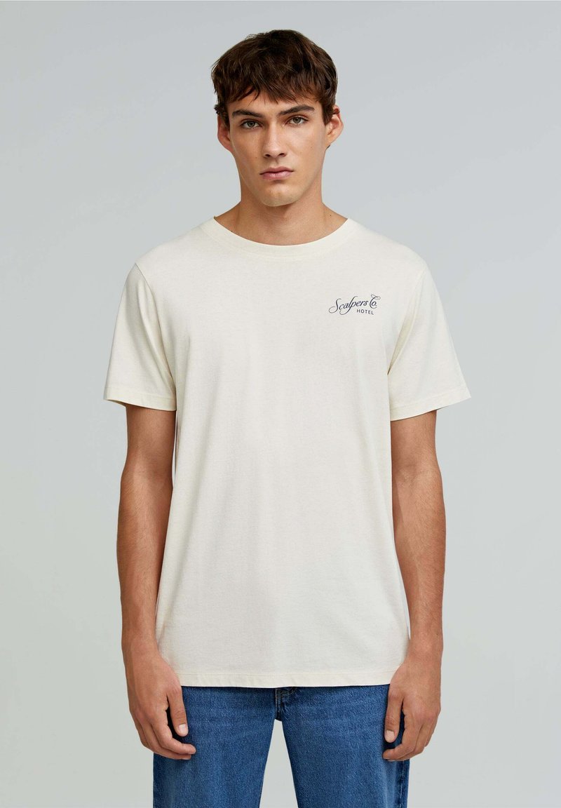 Cremefarvet bomulds t-shirt med korte ærmer, der har et lille marineblåt logo på venstre bryst og en klassisk rund halsudskæring.