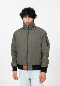 Veste bomber vert olive avec un col montant, une fermeture éclair, des poignets côtelés et une poche zippée sur la manche ; elle est dotée d'une étiquette orange à l'ourlet.