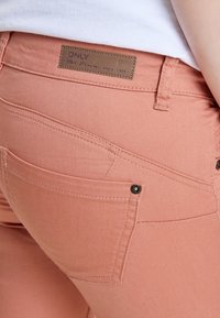 Los jeans de denim rosa presentan un parche con logo, acentos de botones y un diseño de bolsillo estructurado. La tela es suave y tiene un ligero estiramiento.