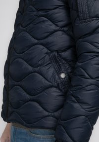 Marineblauer gesteppter Pufferjacke mit Reißverschluss, sichtbaren Nähten und einer Seitentasche mit silberner Knopfdetaillierung. Texturierte Oberfläche des Materials.