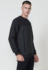 Sudadera negra con parte superior texturizada y paneles inferiores lisos, con un diseño de texto impreso. Mangas largas y puños acanalados.