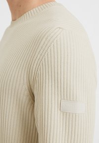 Beige gerippter Pullover mit rundem Halsausschnitt und strukturiertem Stoff. Verfügt über ein Markenzeichen auf der linken Schulter als Detail.