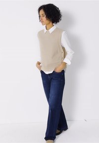 Béžová pletená vesta přes bílou košilovou košili, v kombinaci s tmavě modrými rozšířenými džíny. Vesta má žebrovaný lem a boční knoflíky.