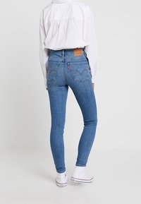 Jeans ajustados de color azul hechos de mezclilla, con una cintura alta, bolsillos traseros y patrones de costura distintivos. Combinados con zapatillas blancas.