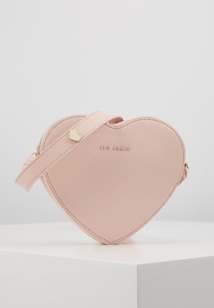 Petit sac bandoulière en cuir rose clair en forme de cœur avec logo Ted Baker doré et bandoulière réglable sur une surface blanche.