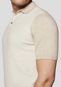 Koszulka polo z krótkim rękawem w jasnym beżu z fakturowanym wzorem w kratę. Posiada klasyczny kołnierzyk i dwukierunkowe zapięcie na guziki. Miękka, dzianinowa tkanina.