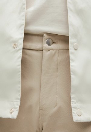Pantalon beige clair avec une texture lisse et une fermeture à bouton métallique. Associé à une veste blanche légère avec des boutons pression.