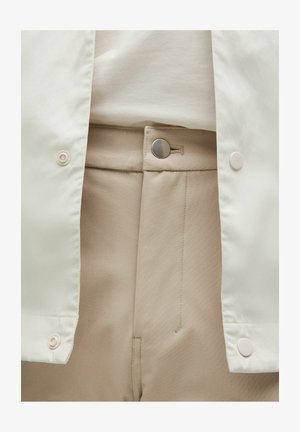 Pantalon beige clair avec une texture lisse et une fermeture à bouton métallique. Associé à une veste blanche légère avec des boutons pression.