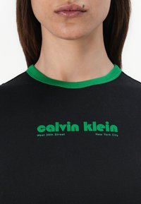 Black T-shirt with green ribbed collar. Features "calvin klein" and "West 39th Street, New York City" printed in bold green lettering.