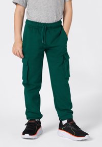 Enfant portant un pantalon de survêtement cargo vert, un t-shirt gris et des baskets noires avec des semelles blanches et des accents orange, se tenant avec une main dans la poche.