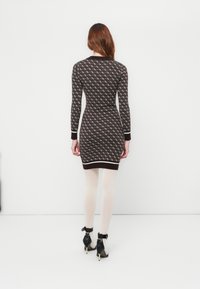 Una donna con i capelli lunghi indossa un vestito scuro aderente e decorato, collant bianchi e scarpe con tacco a jof black e cinturini alla caviglia, front facing a white wall.