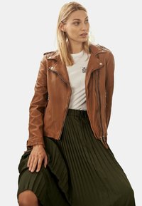 Braune Leder-Bikerjacke mit silbernen Reißverschlüssen, getragen über einem weißen T-Shirt, kombiniert mit einem dunkelgrünen, gefalteten Rock.