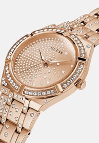 Rožnato zlato uro s okroglo številčnico, ki se ponaša s kristali okrašenim kazalcem in okvirjem. Ravna kovinska zapestnica s poliranim zaključkom in nastavljivimi povezavami.