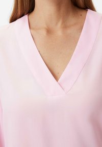 Blouse rose clair avec un col en V, en tissu lisse, présentant une légère brillance. Le design est minimaliste, sans motifs ni accents.