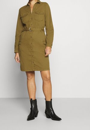 Femme portant une robe boutonnée vert olive jusqu'aux genoux avec poches et ceinture, associée à des bottines noires à talons.