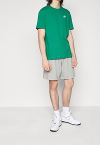 Grön bomulls-t-shirt med korta ärmar och liten vit logga, tillsammans med ljusgrå shorts och vita sneakers med svarta detaljer.