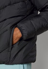 Main droite insérée dans la poche d'une doudoune noire portée par-dessus un pantalon bleu clair, sur fond gris.