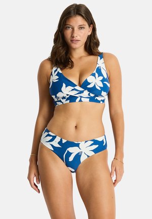 Vrouw draagt een blauwe bikini met grote witte bloemenprint, staat en kijkt recht vooruit tegen een effen lichte achtergrond.