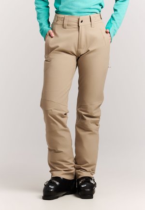 Personne portant un pantalon de ski beige, une veste turquoise texturée et des chaussures de ski noires, debout les mains dans les poches.