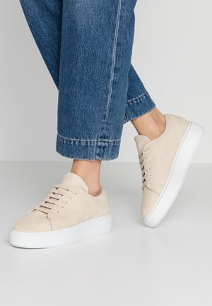 Osoba nosząca jasnobeżowe zamszowe sneakersy z białą grubą podeszwą i niebieskie jeansy na jednolitym szarym tle.