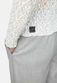 Weißes, langärmeliges Spitzenoberteil mit filigranen Mustern und gewelltem Saum, kombiniert mit hellgrauen, maßgeschneiderten Hosen mit seitlichen Taschen.