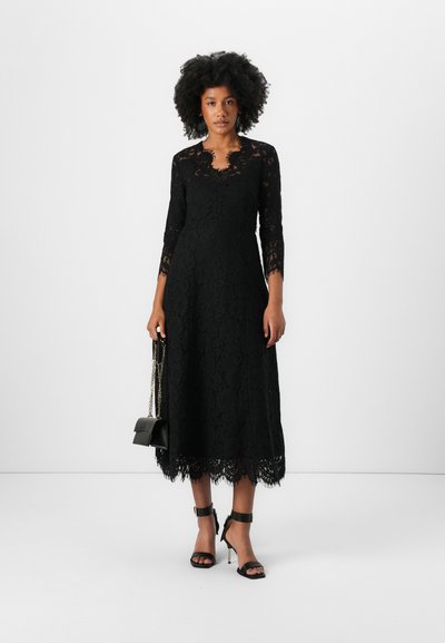 Robe midi en dentelle noire avec des manches trois-quarts, un décolleté en forme de vagues et un ourlet détaillé. Le modèle porte un petit sac à main noir et des talons noirs.