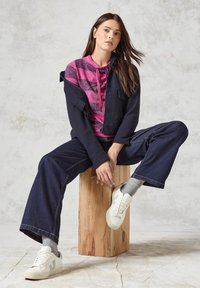 Veste zippée marine sur un hoodie rose à motifs, associée à un pantalon large en jean noir et des baskets blanches avec des accents verts. Fond en tabouret en bois.