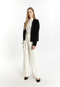 Zwarte cropped cardigan over een crèmekleurige blouse, gecombineerd met hoog-getailleerde, wijde witte broeken en zwarte puntige hakken.
